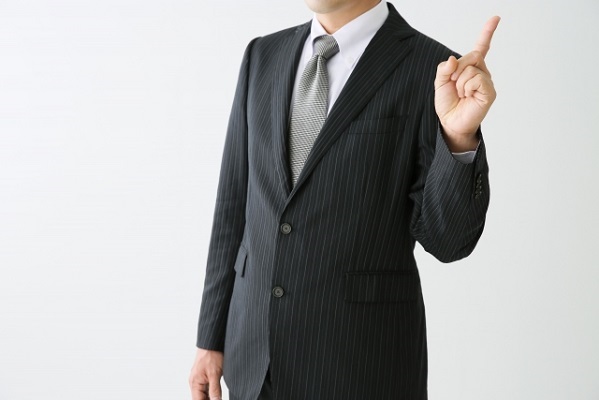 左手の人差し指を立てているスーツの男性の写真
