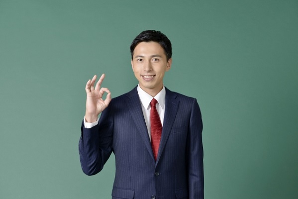 緑の背景に右手で丸を作って笑顔のスーツ姿の男性の写真
